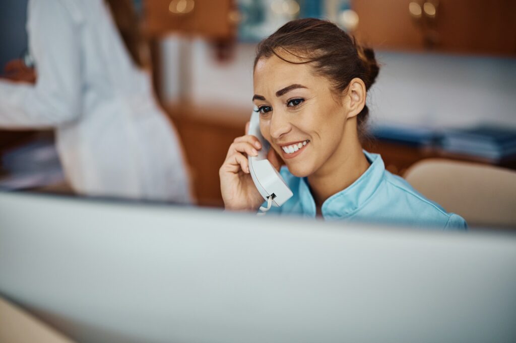 Happy nurse talking on the phone while working on a computer at medical clinic.