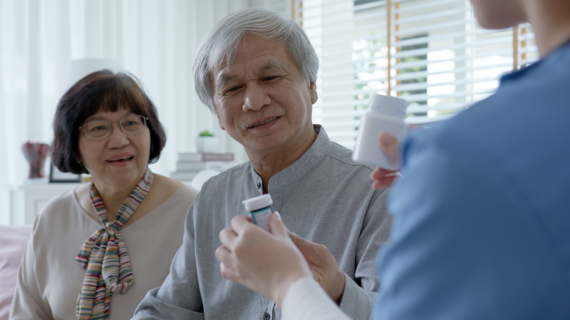 Young caregiver in scrubs uniform showing medicine bottle to elderly asian couple